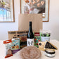 A curated Heritage Day food gift set arranged on a white table in front of a brown paper bag. The set includes a large round of cured meat, a bottle of wine, a can of almond oil, a bag of pistachios, a package of figs, a wedge of cheese, and a small jar, with branding visible on several items.