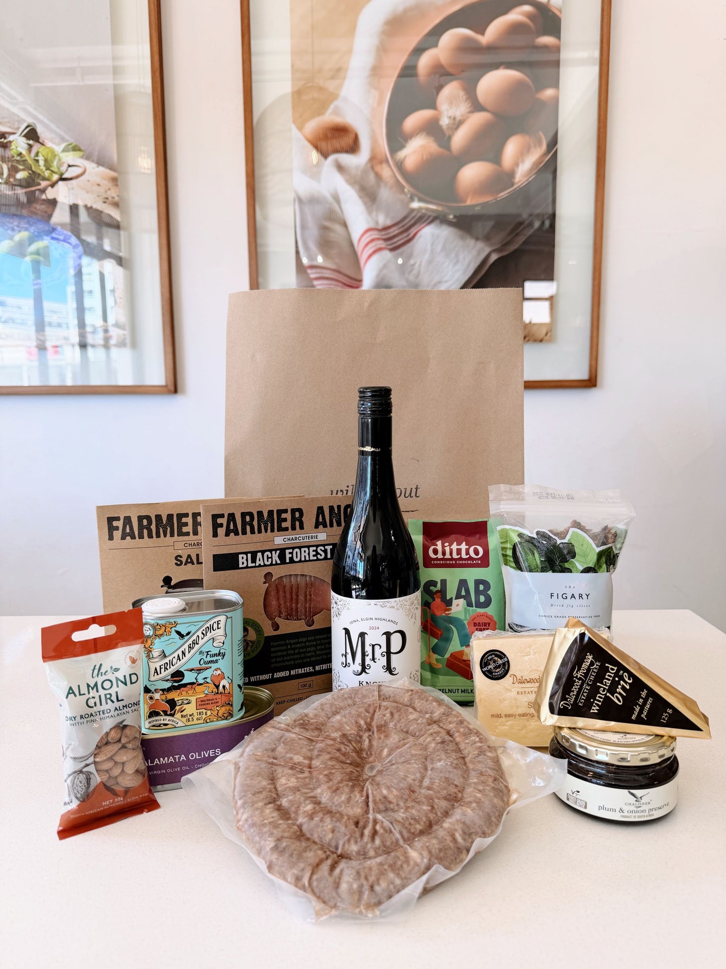 A curated Heritage Day food gift set arranged on a white table in front of a brown paper bag. The set includes a large round of cured meat, a bottle of wine, a can of almond oil, a bag of pistachios, a package of figs, a wedge of cheese, and a small jar, with branding visible on several items.