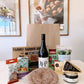 A curated Heritage Day gift basket on a white table, featuring a large brown paper bag, a bottle of M&P wine, a bag of Dittlo salad, artisanal cheeses, crackers, and a round of cured meat, set against a background of framed wall art.