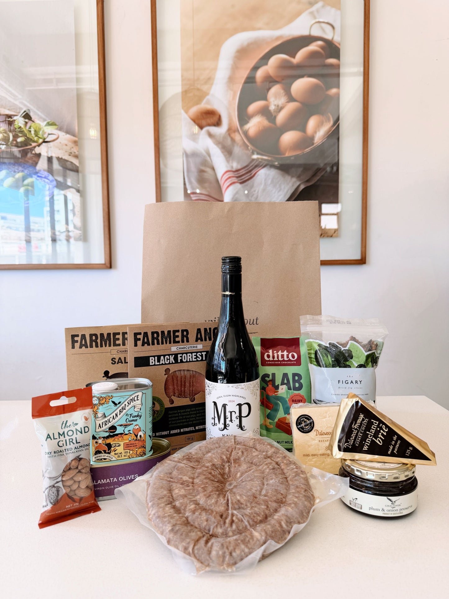 A curated Heritage Day gift basket on a white table, featuring a large brown paper bag, a bottle of M&P wine, a bag of Dittlo salad, artisanal cheeses, crackers, and a round of cured meat, set against a background of framed wall art.